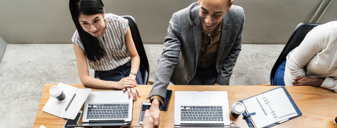 2 people sit in a meeting, one person shakes the hand of a third one over the desk